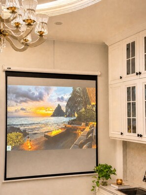 Una habitación con una pantalla de proyector mostrando una escena de atardecer, armarios blancos y una araña.