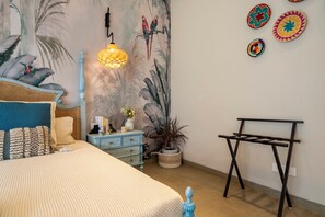 A bedroom with a bed, a nightstand, a dresser, a hanging light, a wall-mounted rack, and a large plant.