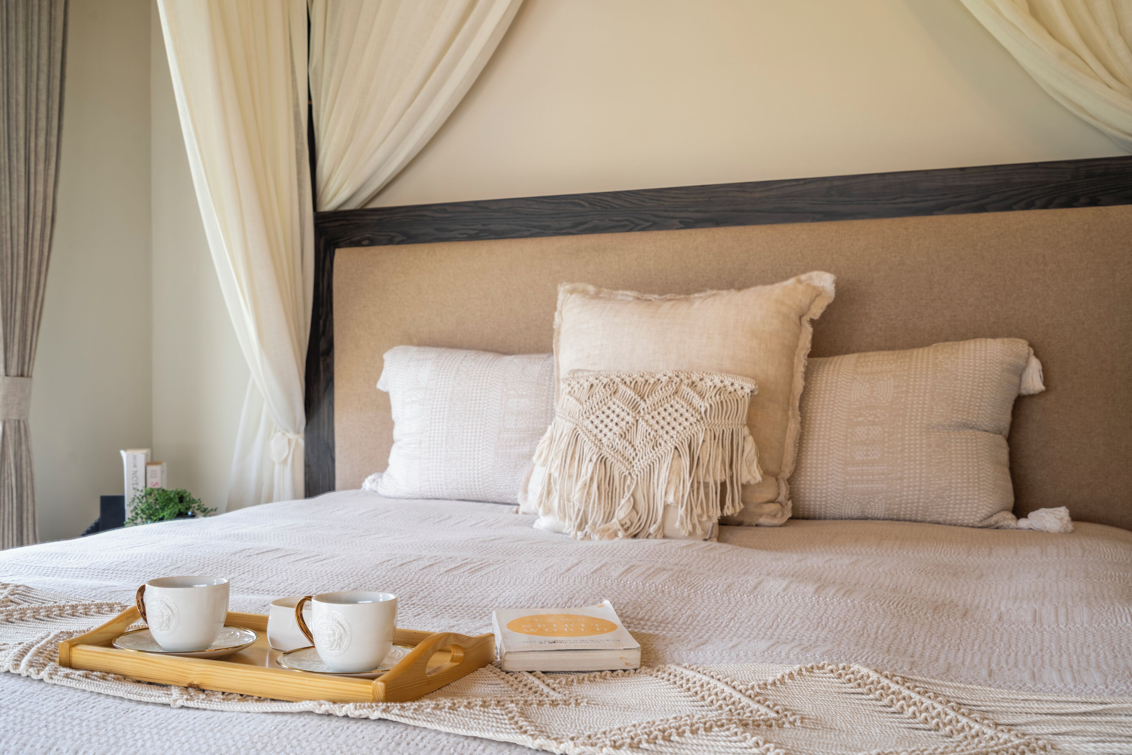 A neatly made bed with a wooden headboard, a tray with cups and a book, and a macrame throw.