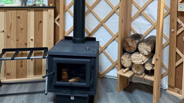 A wood-burning stove with a black exterior, a metal ladder, and a stack of firewood against a wooden lattice wall. - A tranquil yurt with all the amenities in charming Big Lake (Big Lake)