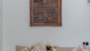 A modern living room with a beige sofa, patterned cushions, a round coffee table, and a rustic wooden wall art piece. - Villa Rame (Uluwatu)