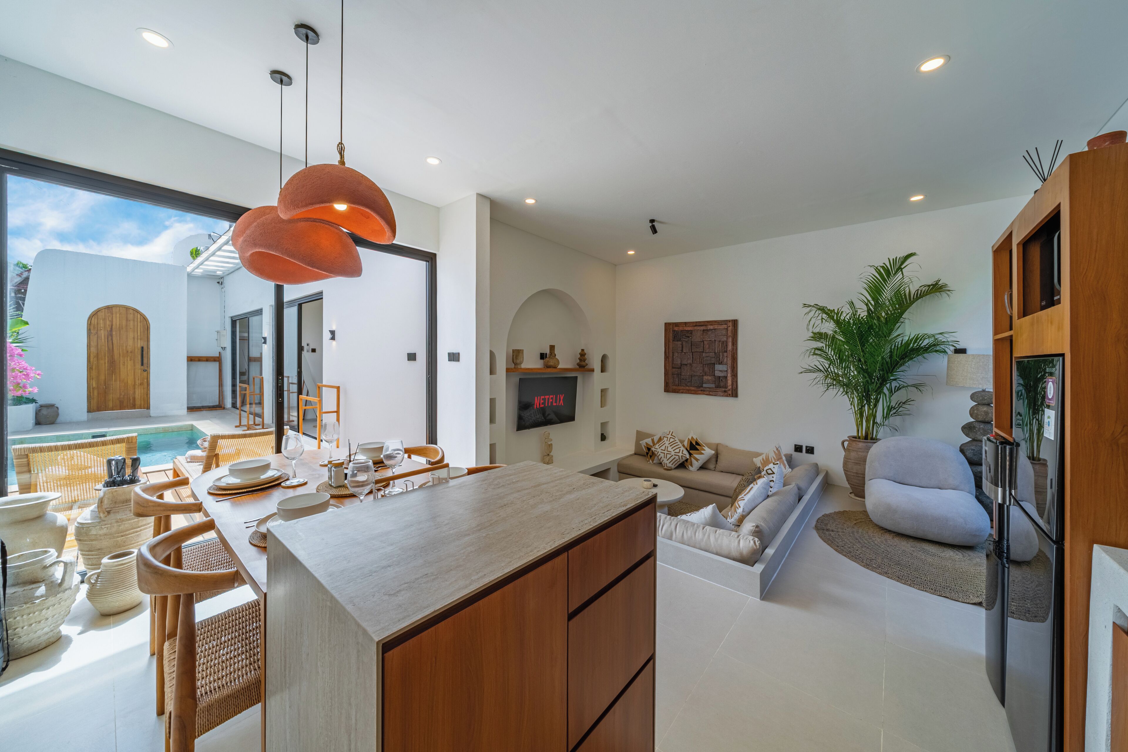 A modern kitchen with a dining area, a living room with a sofa, and a view of the outside.