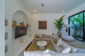 A modern living room with a flat-screen TV, a sofa, a coffee table, and a large window with a view of greenery. - Villa Rame (Uluwatu)