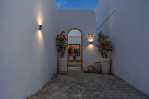 Un sendero estrecho que conduce a la entrada de una villa, con una puerta de madera, dos luces fijadas a la pared y plantas en macetas.