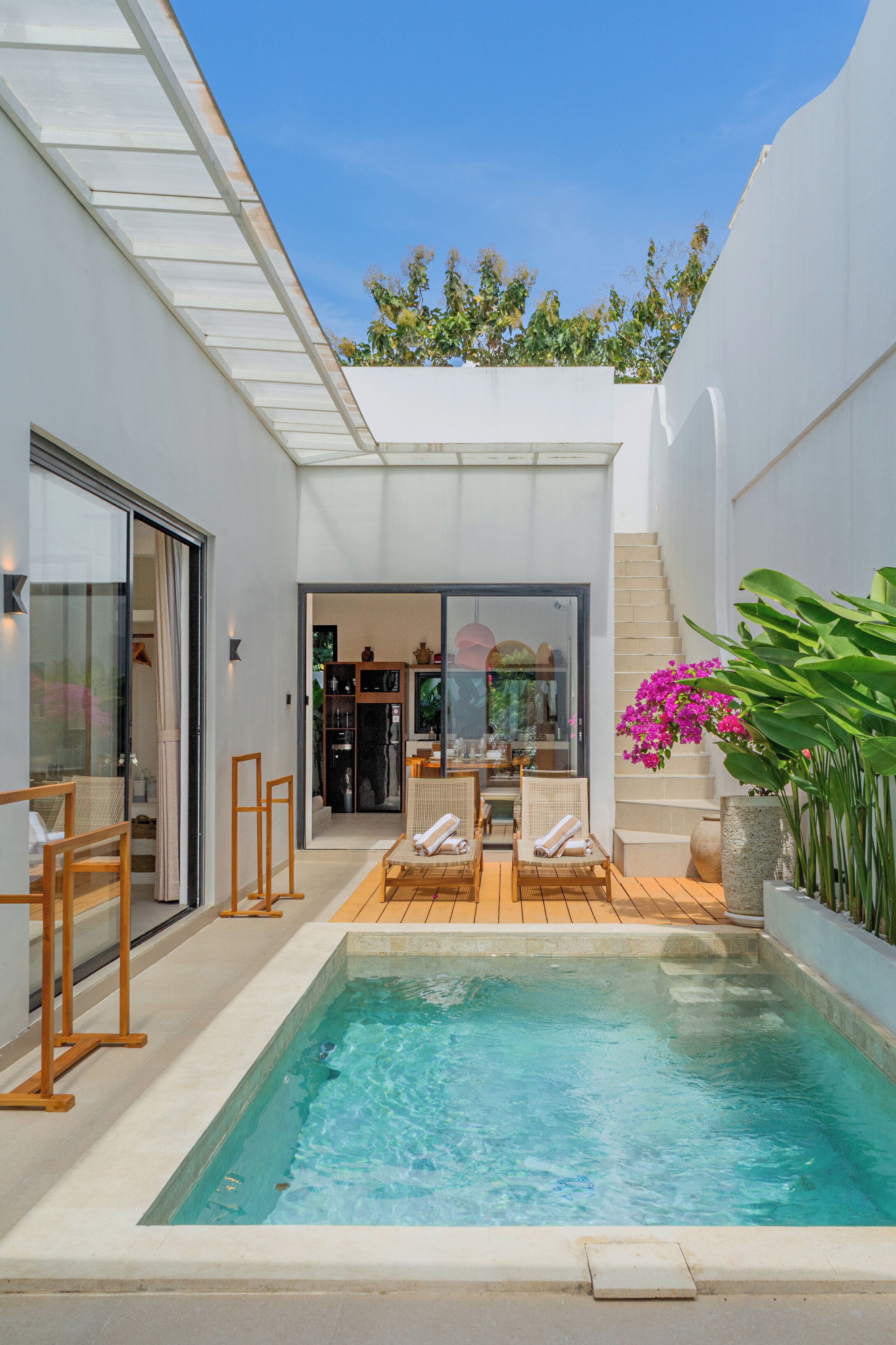 A modern outdoor pool area with a glass door entrance, wooden deck chairs, and a small table.