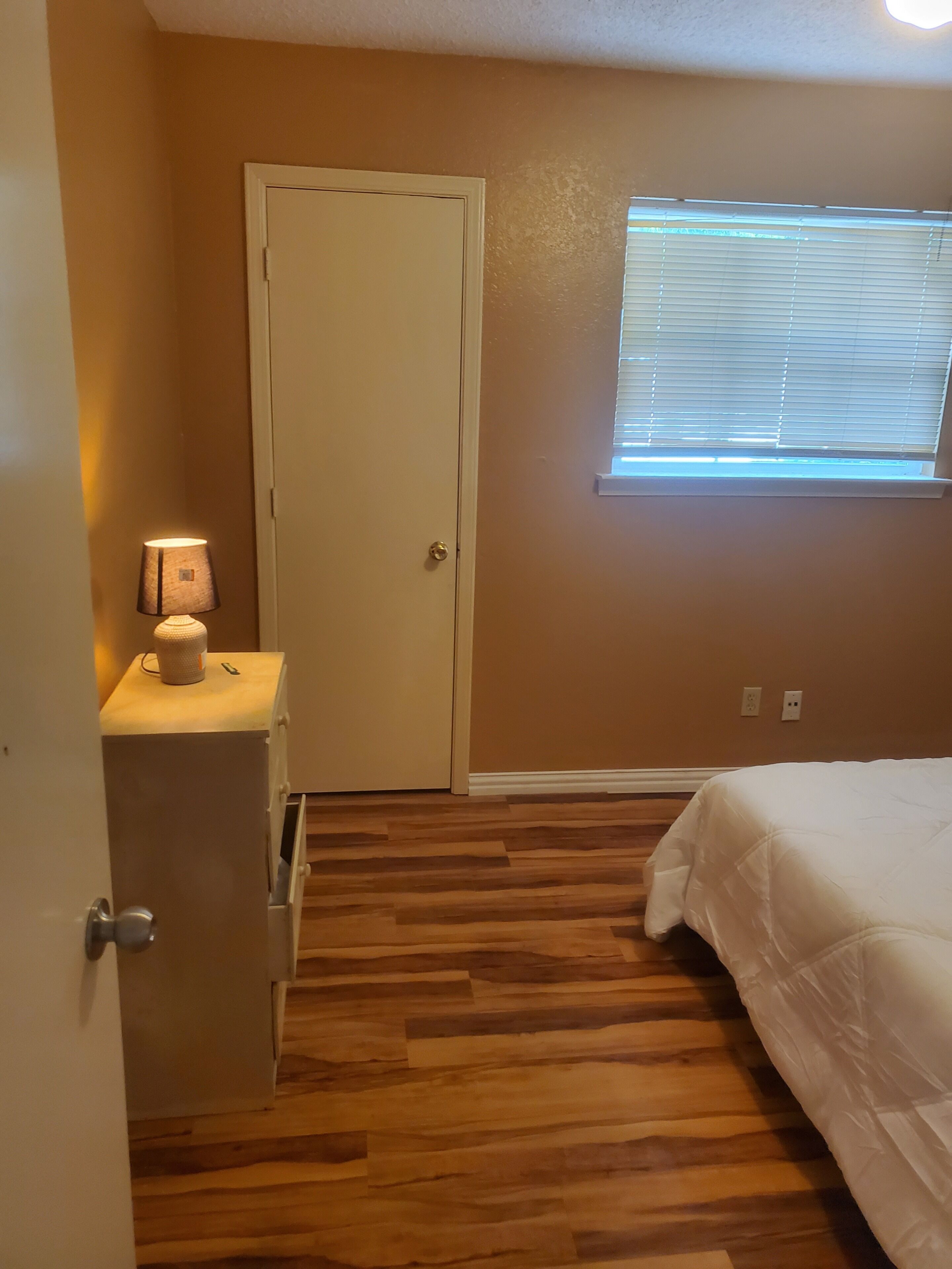 A bedroom with a wooden floor, a bed, a nightstand with a lamp, a window with blinds, and a door.