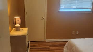 A bedroom with a wooden floor, a bed, a nightstand with a lamp, a window with blinds, and a door.