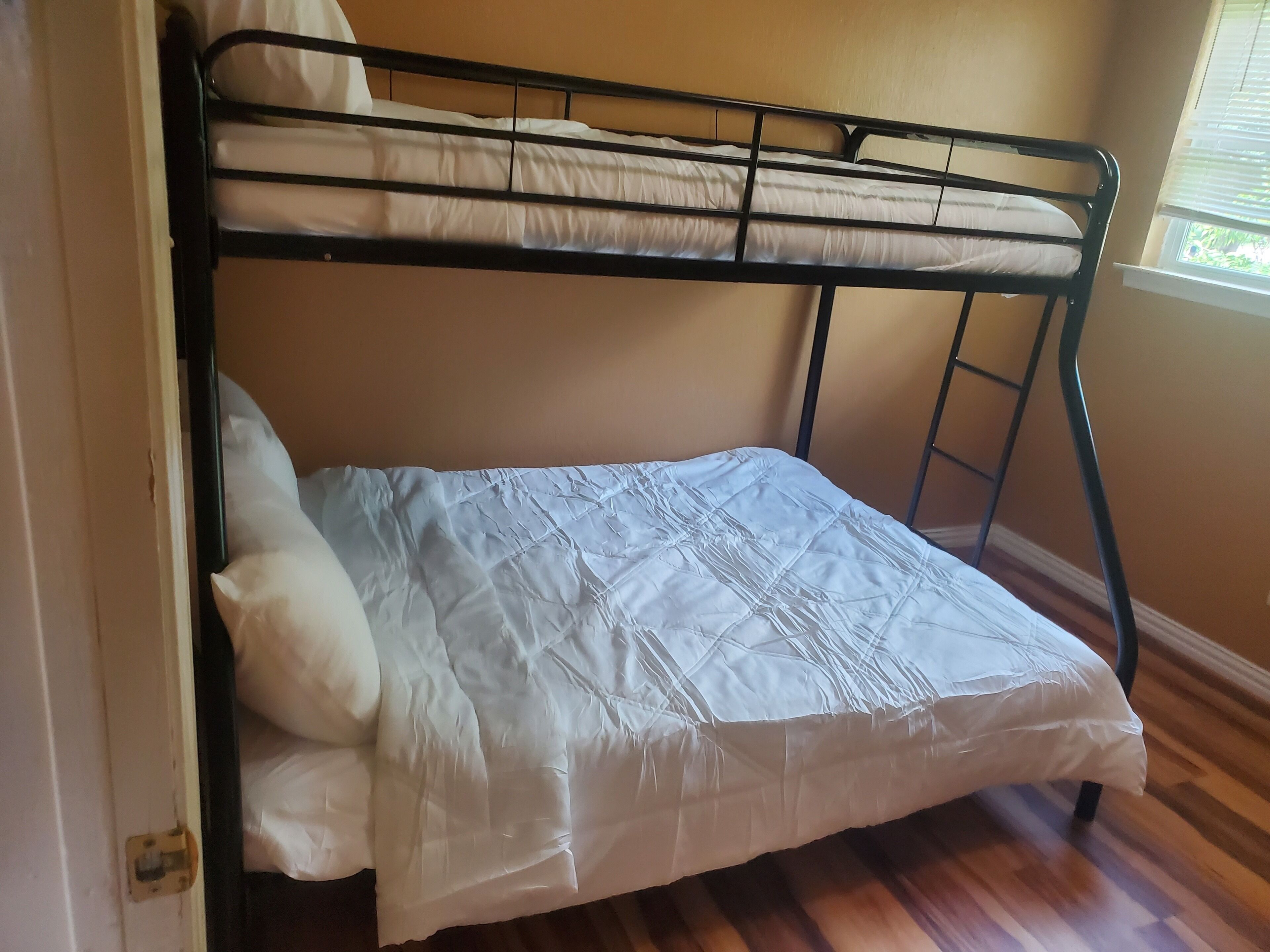 A bunk bed with a mattress and pillows, wooden flooring, and a window with blinds.