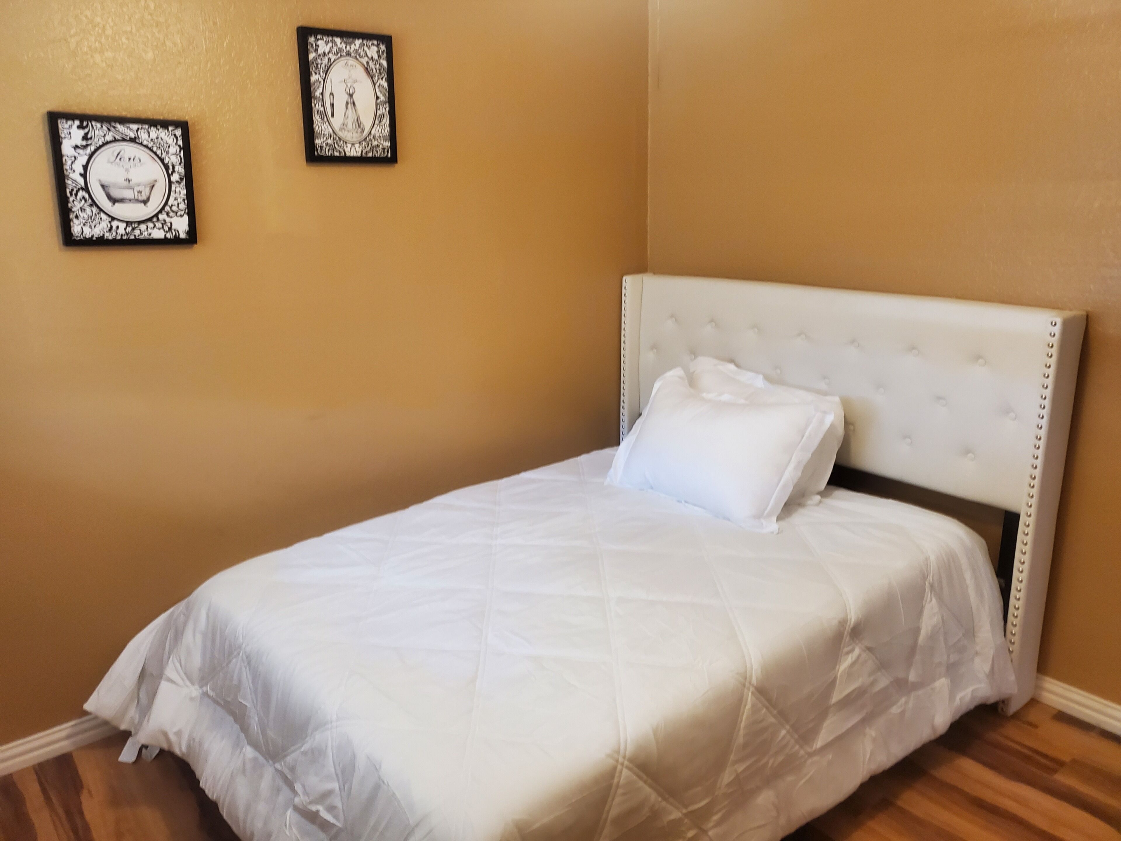 Un lit simple avec une tête de lit et un oreiller blancs, un tableau encadré accroché au mur et un plancher en bois.