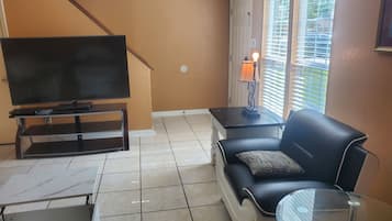 A living room with a flat-screen TV on a stand, a sofa with a cushion, a small table, and a glass coffee table.