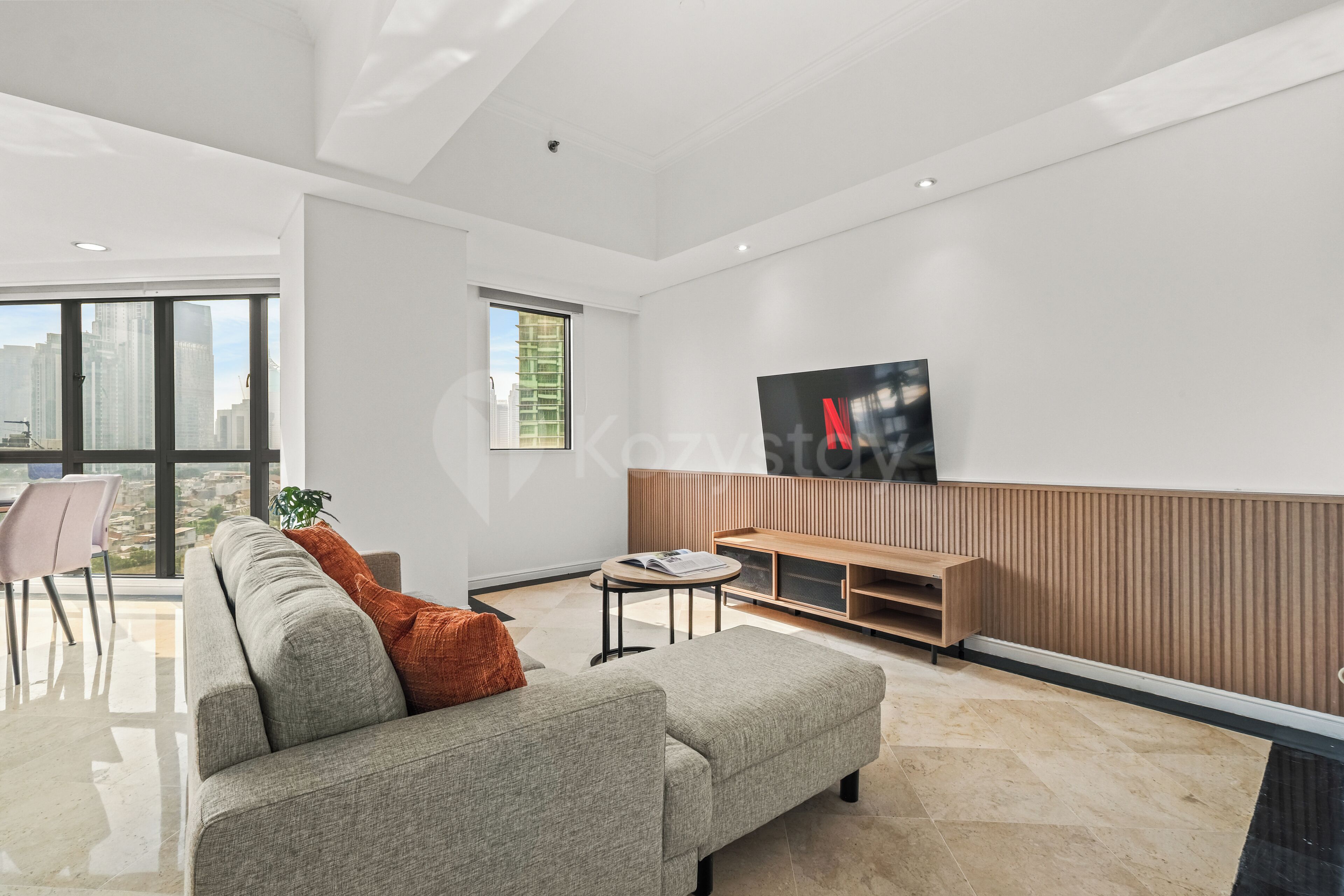 A modern living room with a grey sofa, a coffee table, a TV mounted on the wall, and a window with a view of buildings.