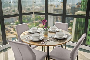 A dining table with four white bowls and plates, set with cutlery, in a room with large windows offering a city view. - Nevora by Kozystay - Kuningan (Jakarta Selatan)