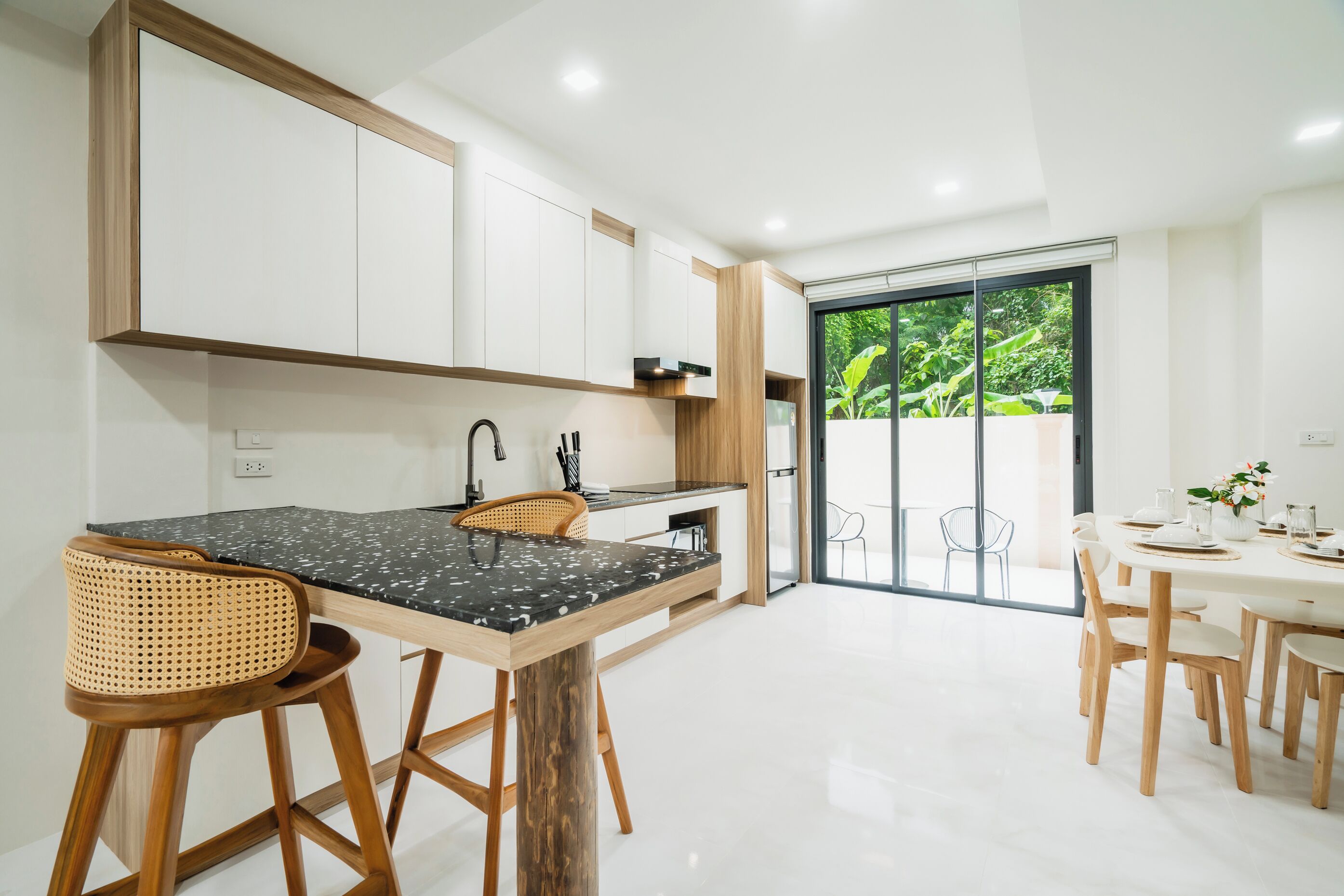 A modern kitchen with a central island, bar stools, a dining area with a white table and chairs, and a sliding glass door leading to a balcony with greenery.