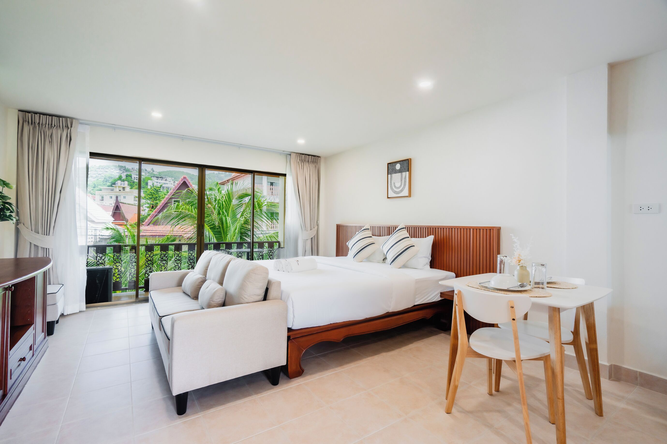 Habitación de hotel con cama, sofá, mesa de comedor con sillas y balcón con vistas a edificios y áreas verdes.