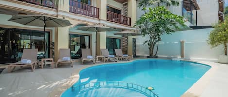 A pool area with lounge chairs and umbrellas, surrounded by a building with multiple balconies and a large tree.