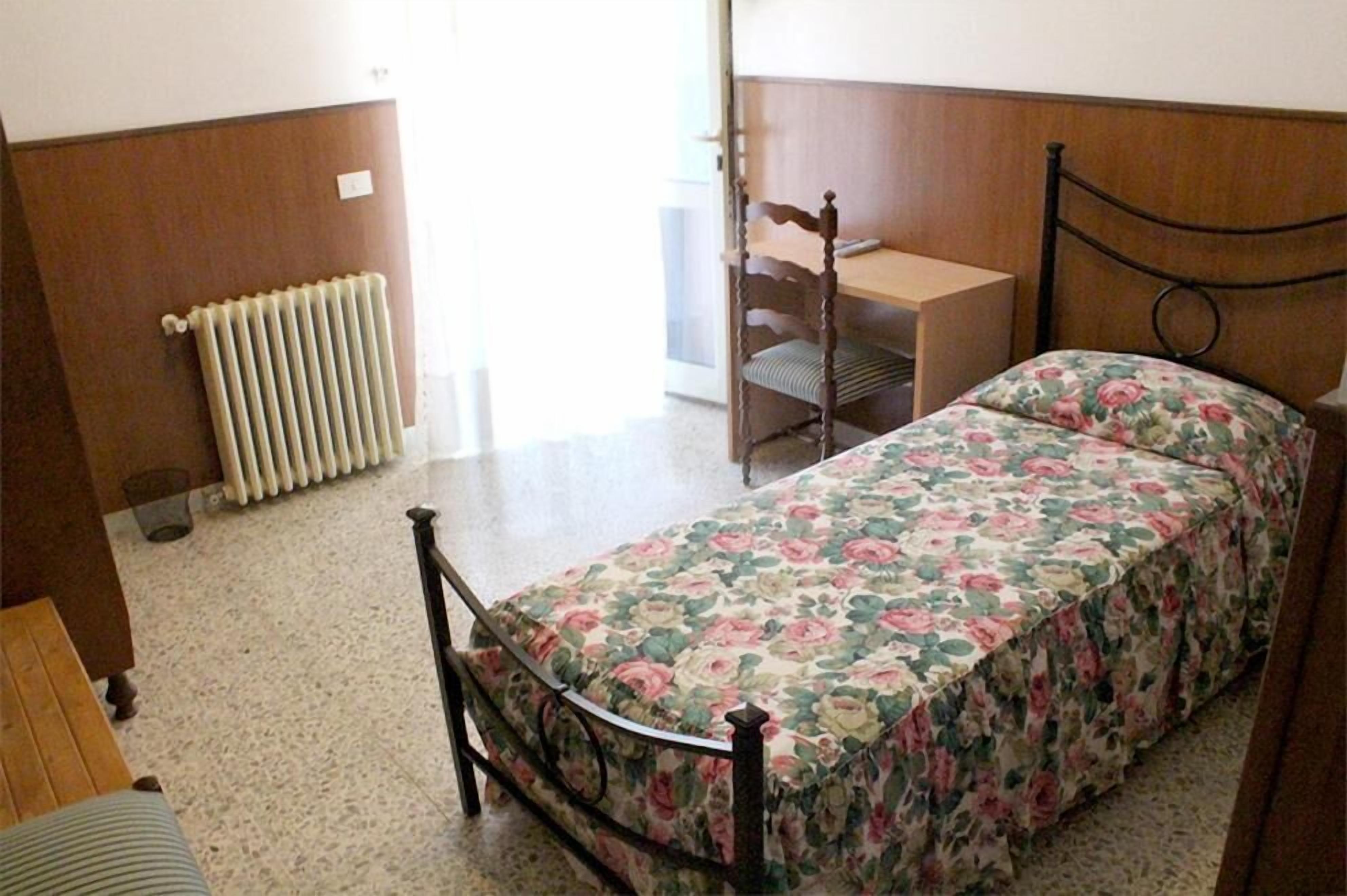 Une petite pièce meublée, comprenant un lit, une chaise en bois, un bureau et un radiateur.