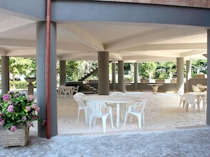Un área de patio al aire libre con sillas y mesas de plástico blanco, rodeada de columnas y un suelo de piedra.