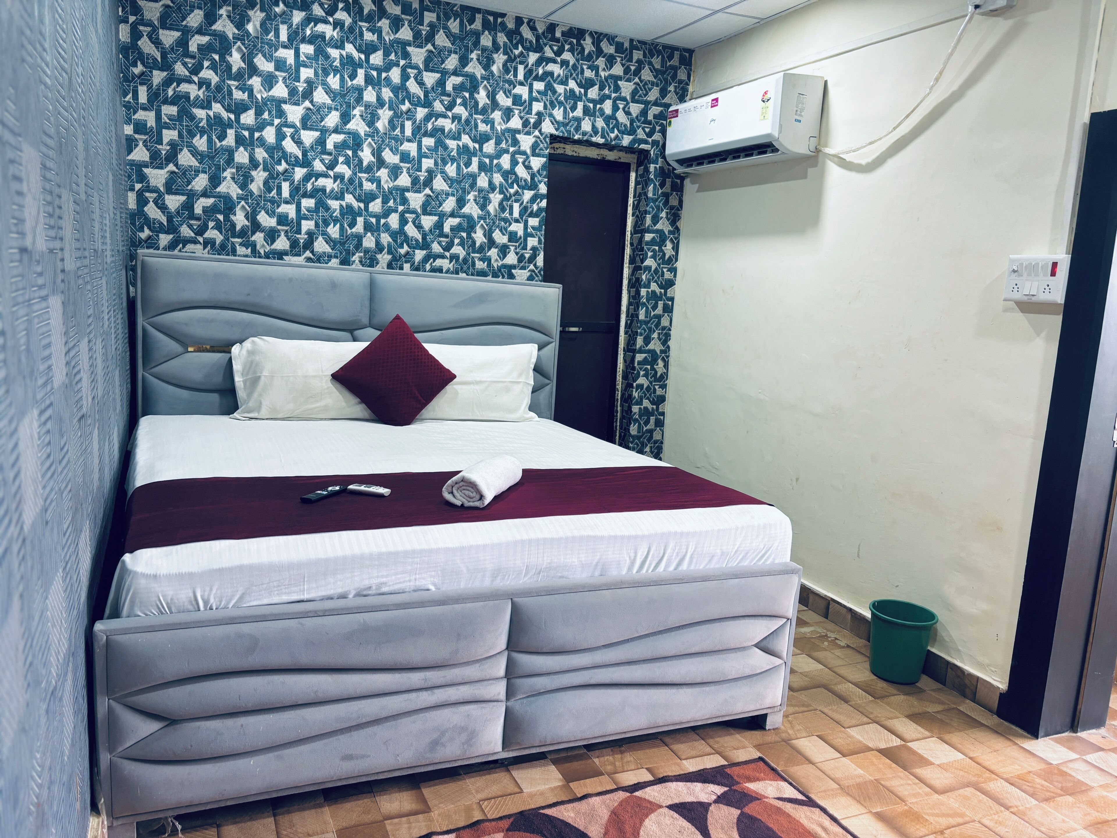 Hotellihuone, jossa on sänky, sängynpääty, kuvioitu seinä, ilmastointilaite, pistorasia ja vihreä roskakori.