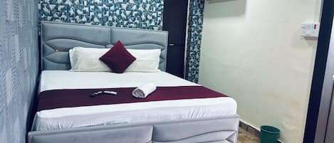 Quarto de hotel com cama, cabeceira, parede estampada, ar condicionado, tomada e lixeira verde.
