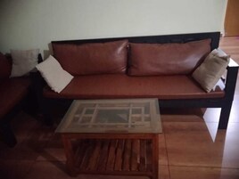 Un sofá de cuero marrón con cojines, una mesa de centro de vidrio y suelo de baldosas.