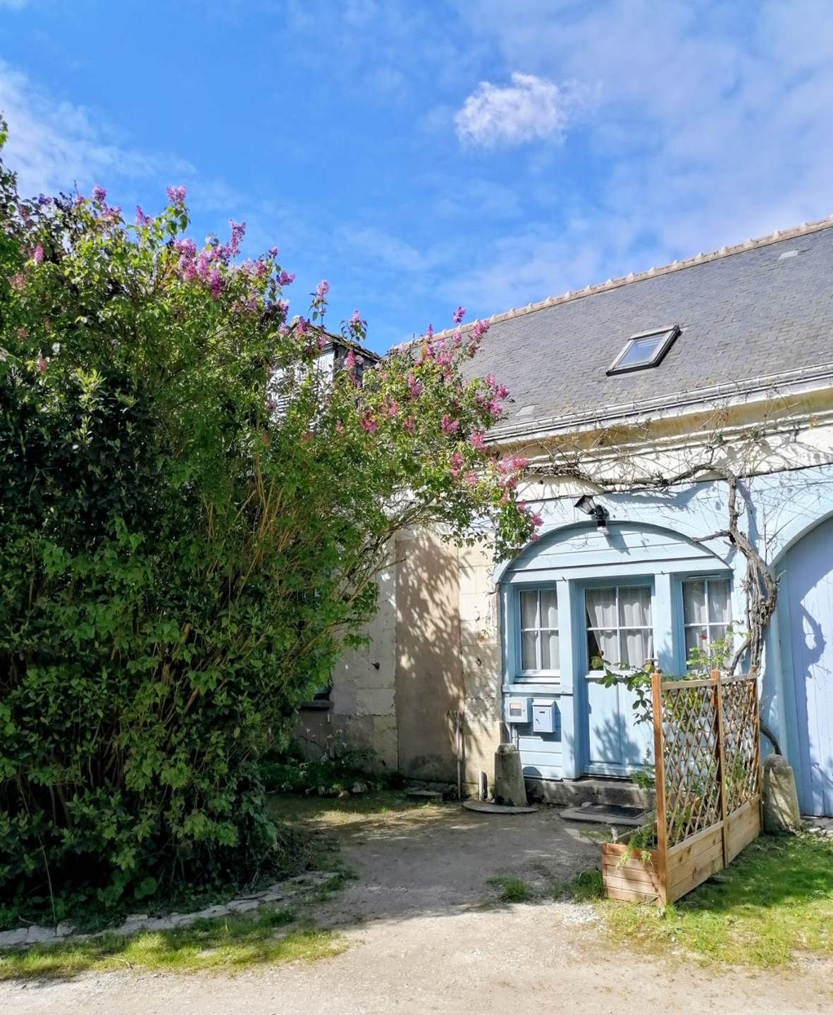 En rustik bygning med en blå facade, en lille veranda og en have med blomstrende planter.