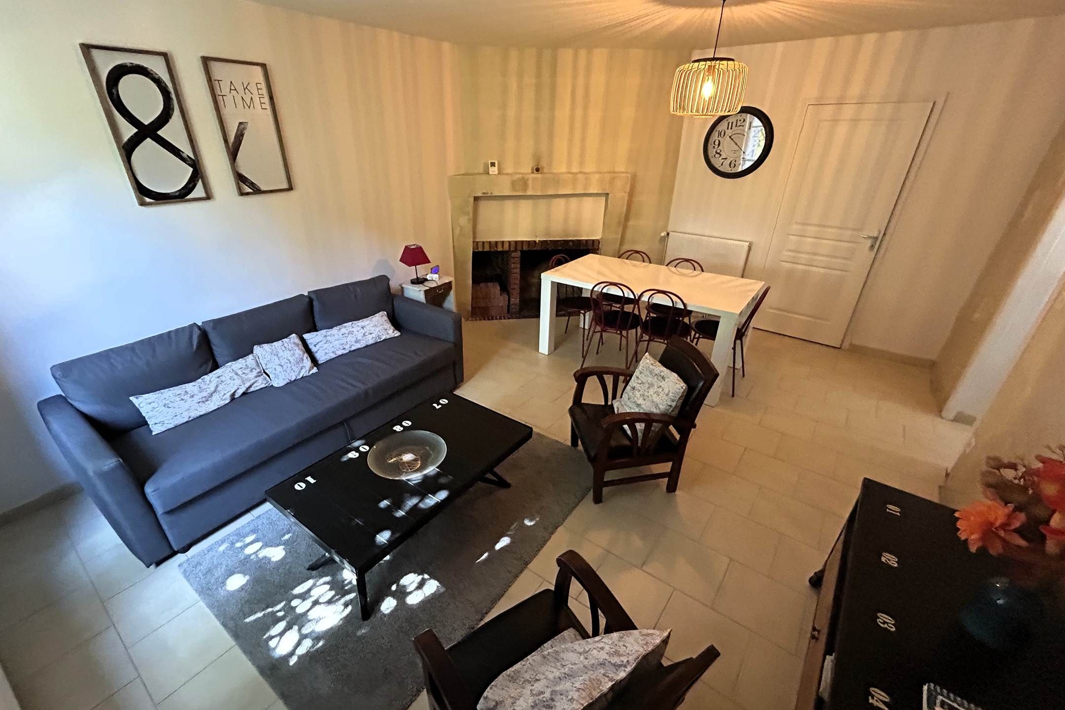 Een woonkamer met een bank, een eettafel met stoelen, een open haard en een klok aan de muur.
