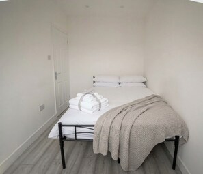 A small, single-bed room with a white bedspread, a beige blanket, and neatly folded white towels. - Brigstock Luxe Stay Modern 2-Bed Apartment (Thornton Heath)