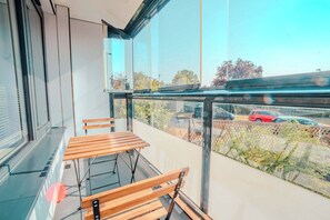 Ein Balkon mit Glastrennwand, Holzmöbeln und Blick auf parkende Autos und Bäume.