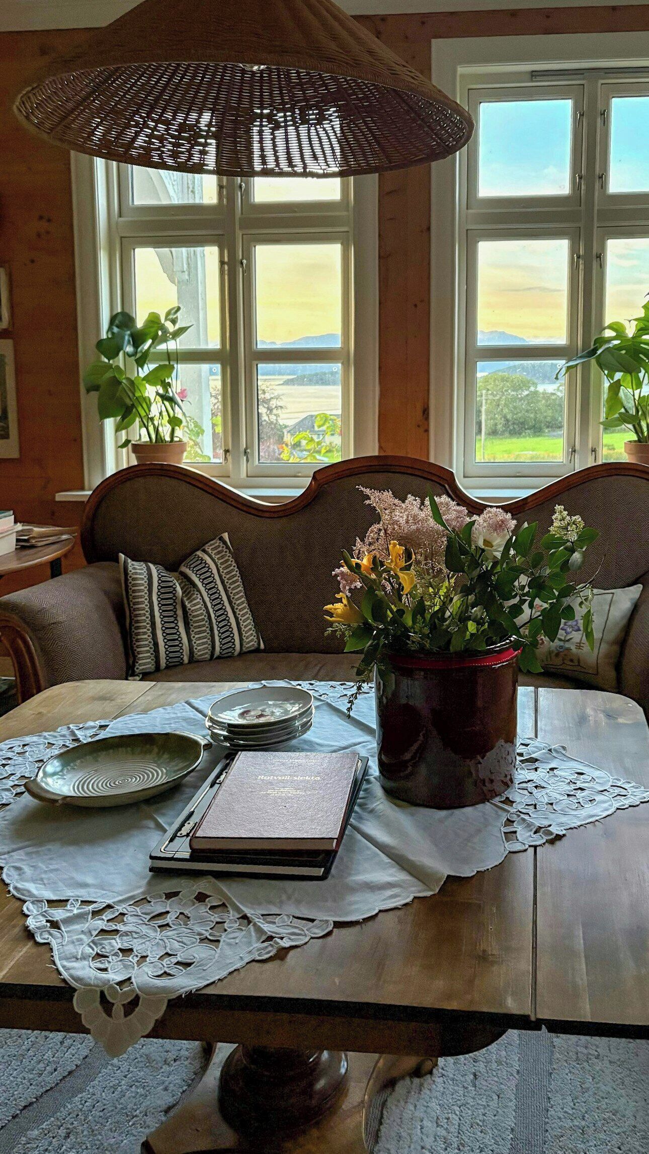 A dining area with a wooden table, a woven umbrella, a vase with flowers, and a view of a landscape through large windows.