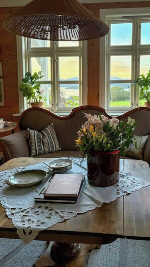 Ein Essbereich mit Holztisch, einem gewobenen Sonnenschirm, einer Vase mit Blumen und Blick auf eine Landschaft durch große Fenster.