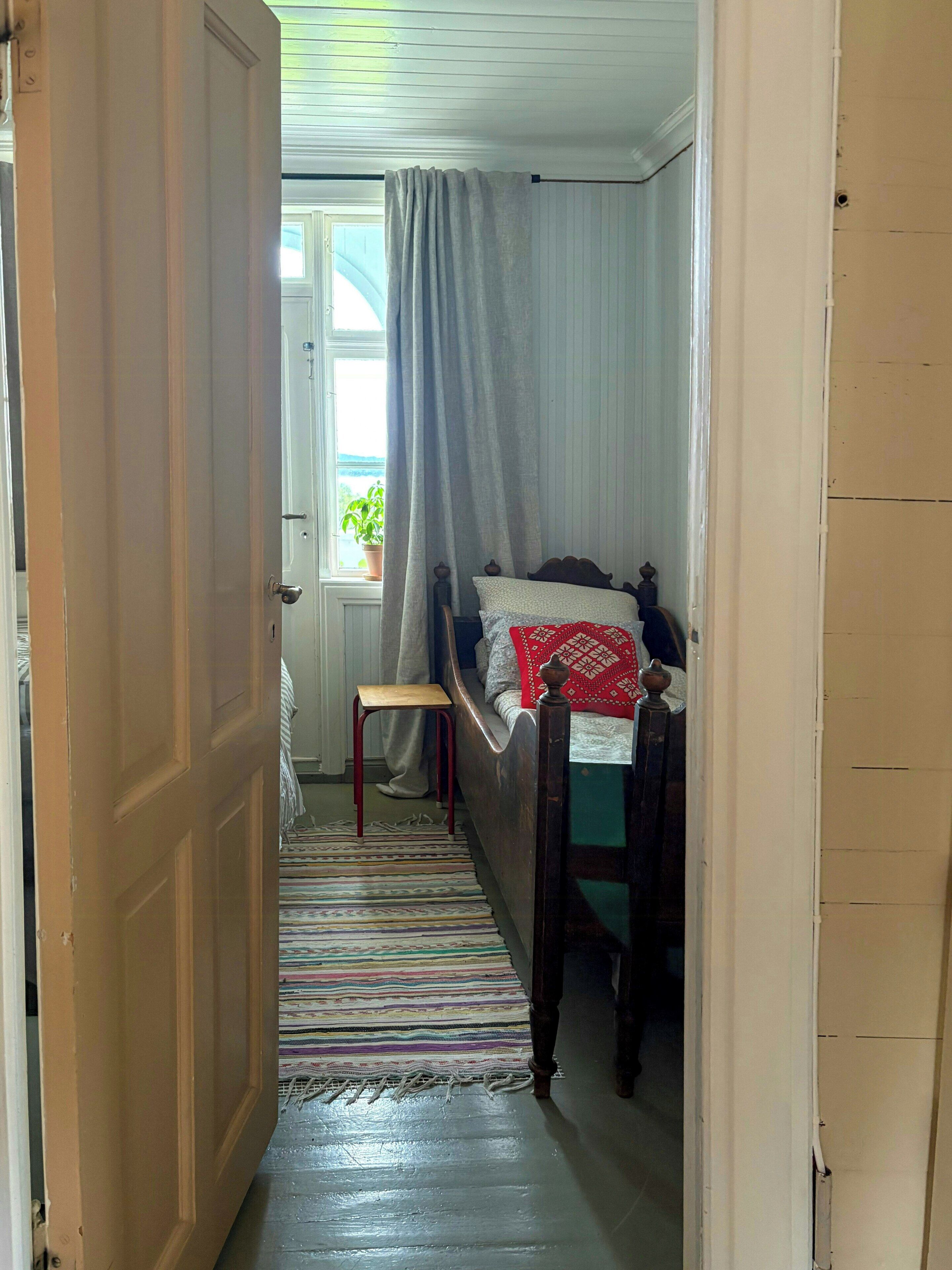 A narrow hallway leading to a bedroom with a bed, a small stool, and a window with curtains.