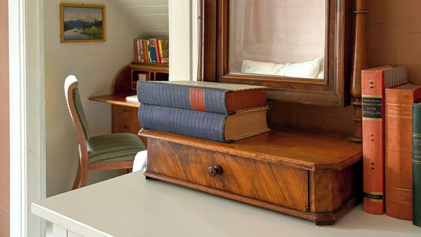 Uma cômoda de madeira com espelho, livros e uma cadeira em um quarto com teto inclinado.