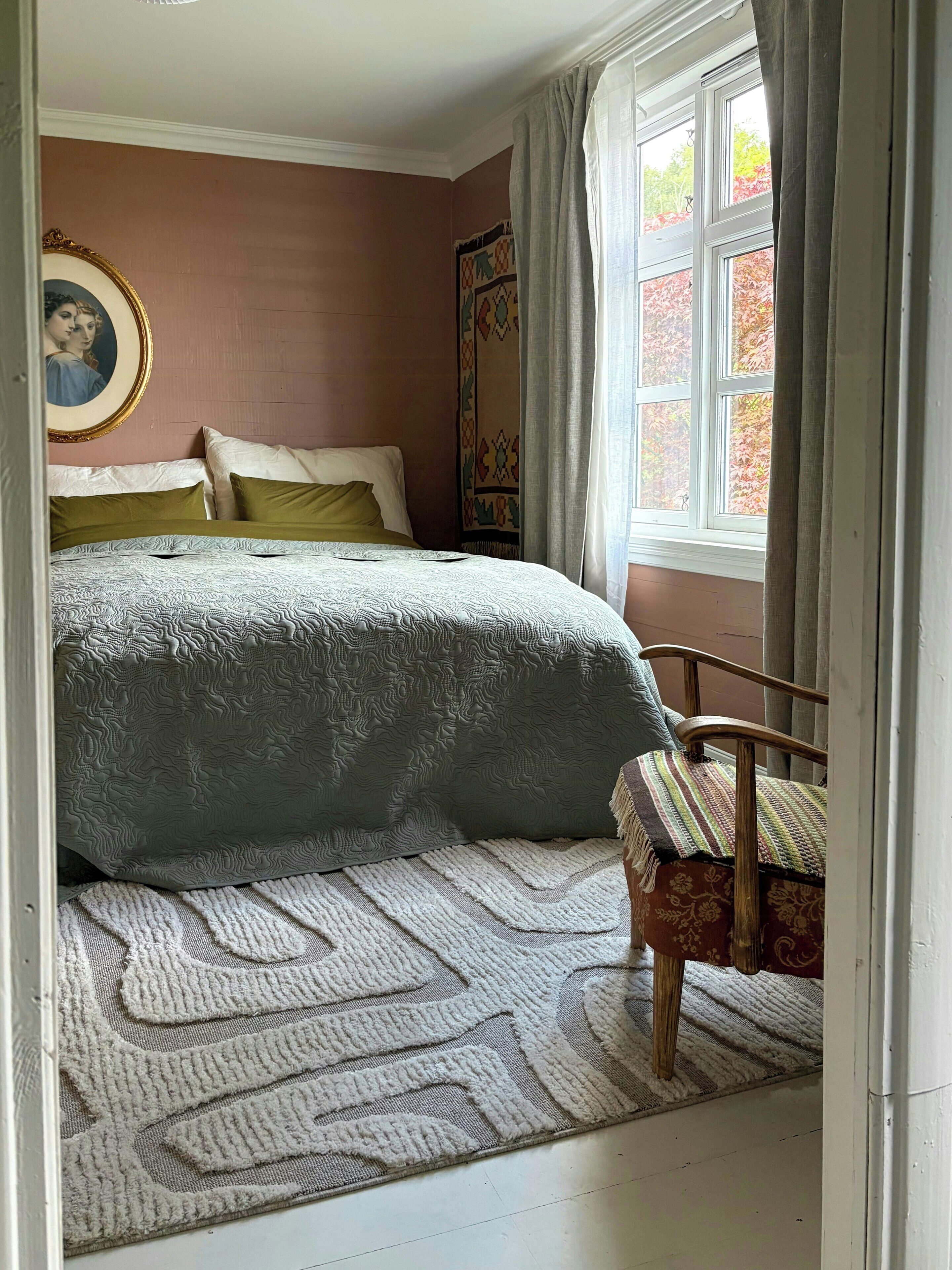 Een slaapkamer met een bed, een stoel, een vloerkleed, een schilderij en een raam met gordijnen.