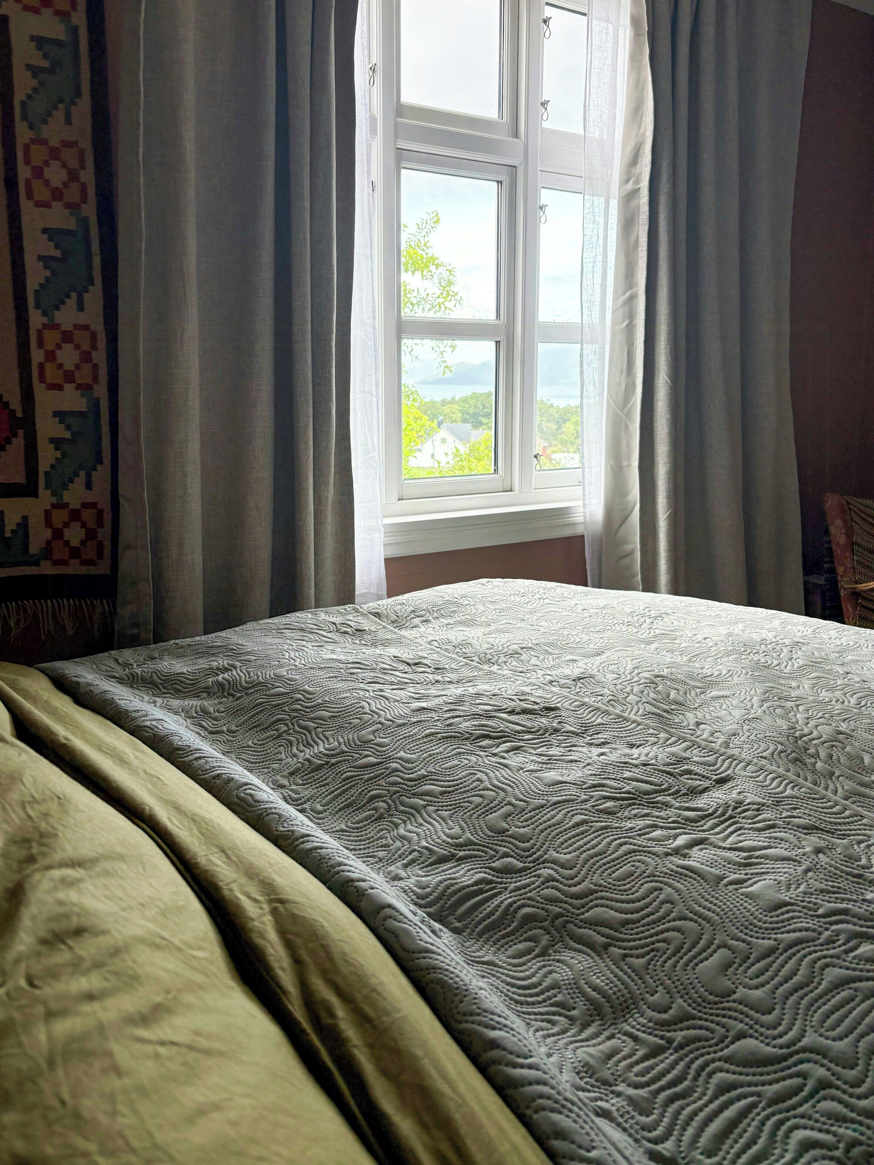 A bed with a patterned bedspread, a window with curtains, and a view of greenery outside.