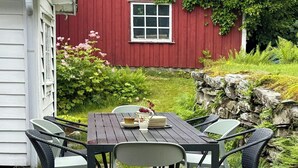 Uma área de refeições ao ar livre com mesa e cadeiras, um edifício vermelho ao fundo e vegetação.