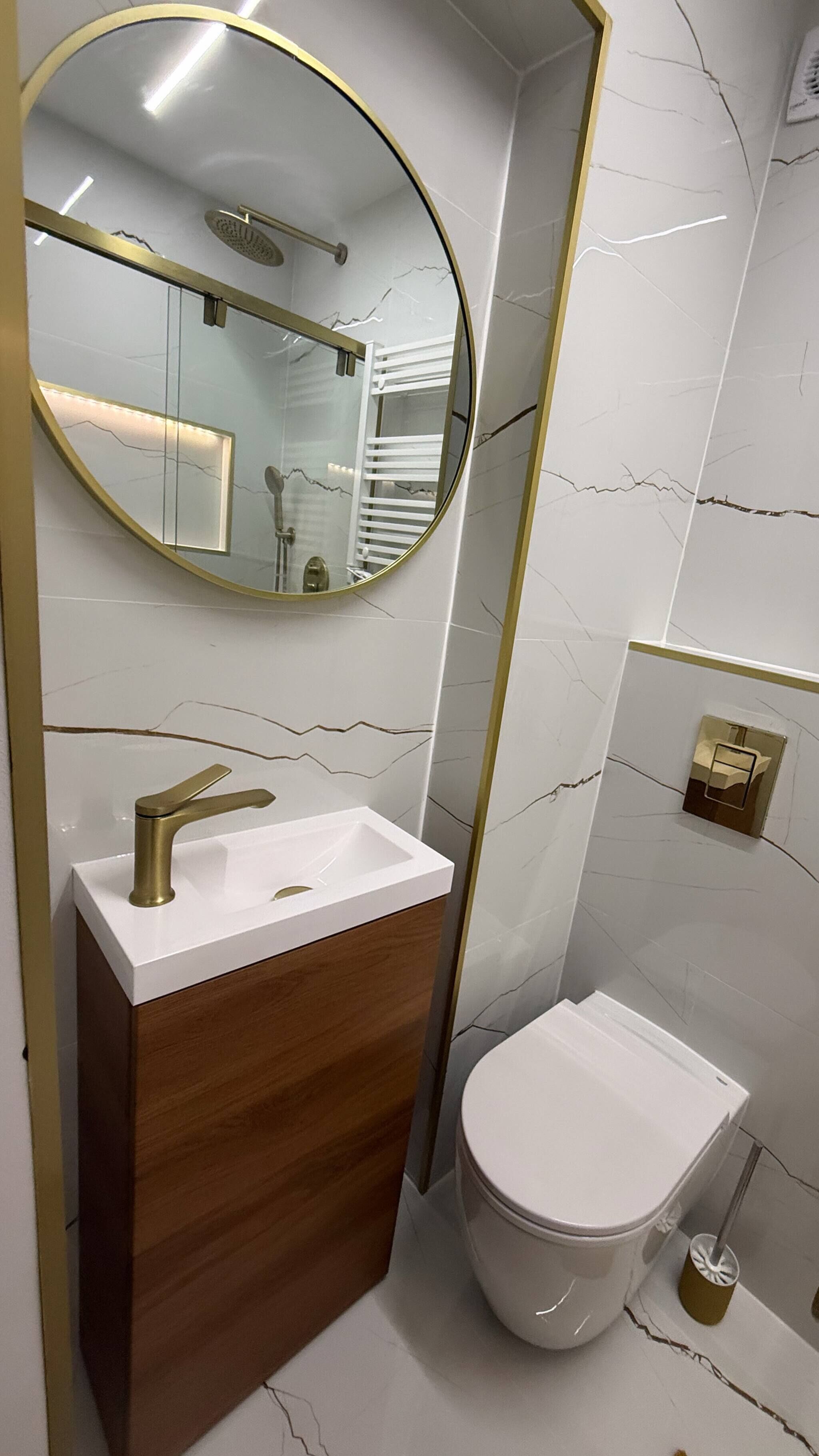 A bathroom with a round mirror, a white sink, a wooden vanity, and a toilet.