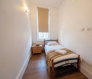 A small, well-lit bedroom with a single bed, a nightstand, a window with blinds, and a wooden floor. - The Luxe Suite - Stylish 3 Bed Apartment (Croydon)
