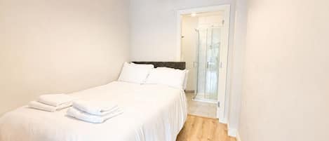 A bedroom with a bed, white linens, a wooden floor, and a bathroom visible through an open door.