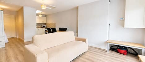 A modern living room with a beige sofa, a wooden floor, a kitchen in the background, and a ceiling light fixture.