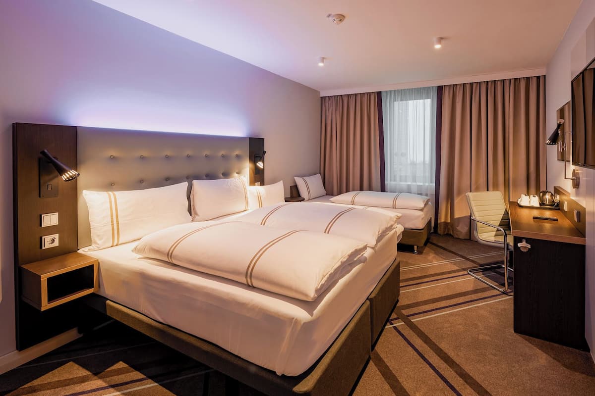 Ein Hotelzimmer mit einem großen Bett, einem kleineren Bett, einem Schreibtisch mit Stuhl, einem Fernseher und einem Fenster mit Vorhängen.