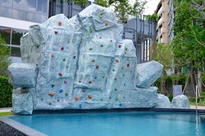 Een klimmuur met kleurrijke grepen, een zwembad en moderne gebouwen op de achtergrond.