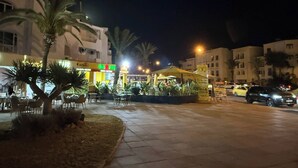 An outdoor dining area with tables and chairs, palm trees, and a building with illuminated signage.