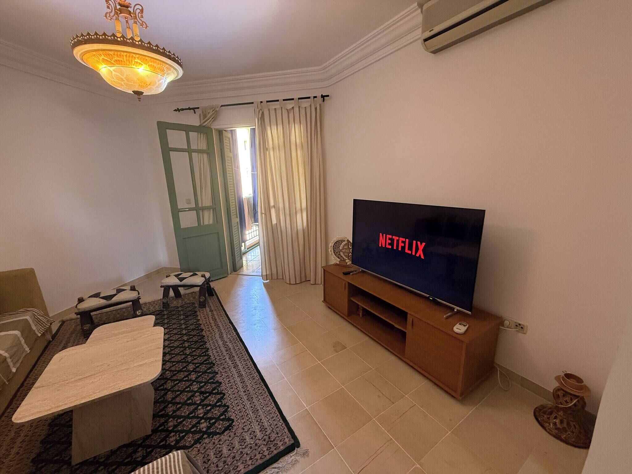 Ein Wohnzimmer mit Fernseher, einer Couch, einem Couchtisch, einem gemusterten Teppich, einer Chandelierleuchtre und einer Tür mit Vorhängen.