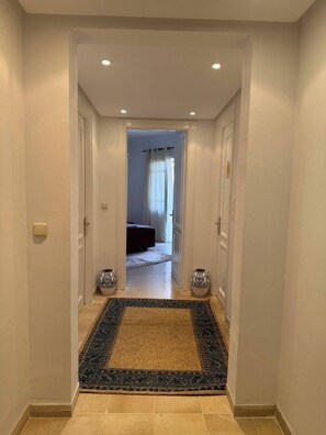 A hallway with a patterned rug, white walls, and a door leading to a room with a bed and curtains.