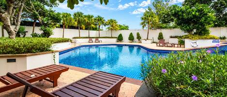 A swimming pool with lounge chairs, surrounded by greenery and a clear sky.