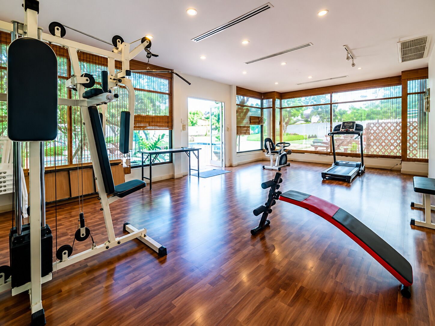 A spacious gym with various exercise equipment, large windows, and a wooden floor.