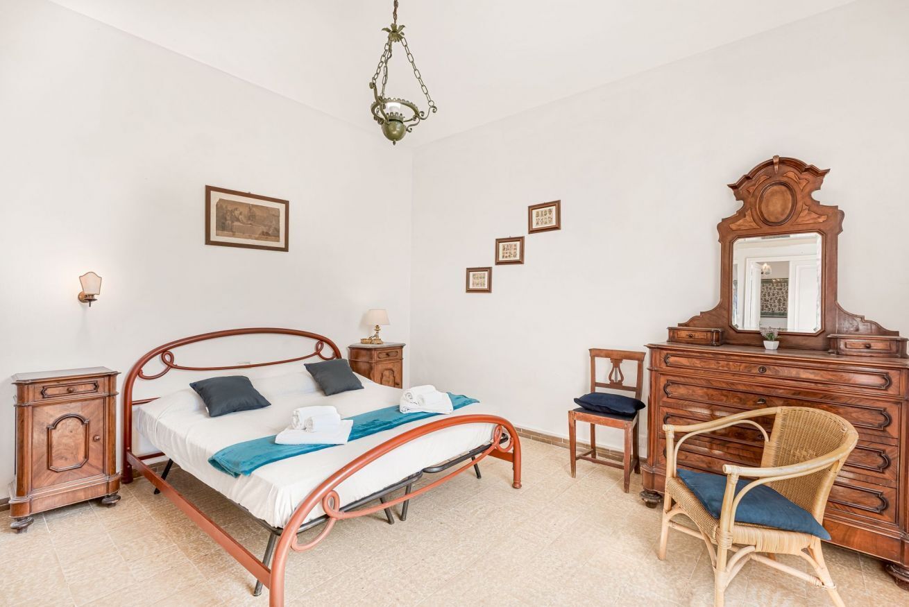 Ein Schlafzimmer mit einem Metallbett, einer Holzkommode, einem Sessel und einer Pendelleuchte.