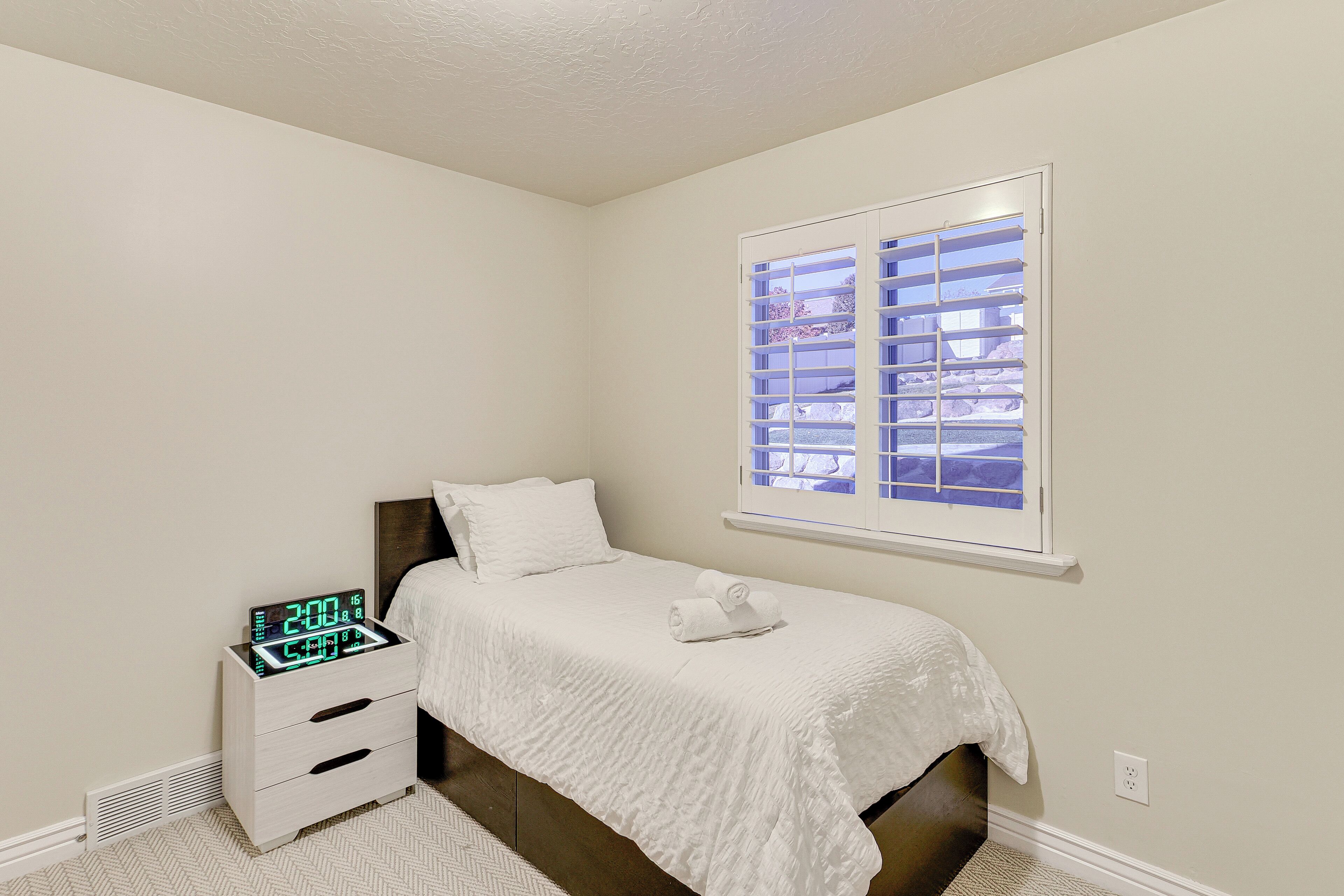 Un dormitorio con una cama, una mesita de noche con un reloj digital, una ventana con persianas y un radiador.
