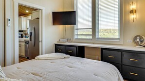A bedroom with a bed, a flat-screen TV mounted on the wall, a desk with a lamp, and a window with blinds.