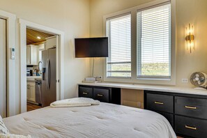 A bedroom with a bed, a flat-screen TV mounted on the wall, a desk with a lamp, and a window with blinds.